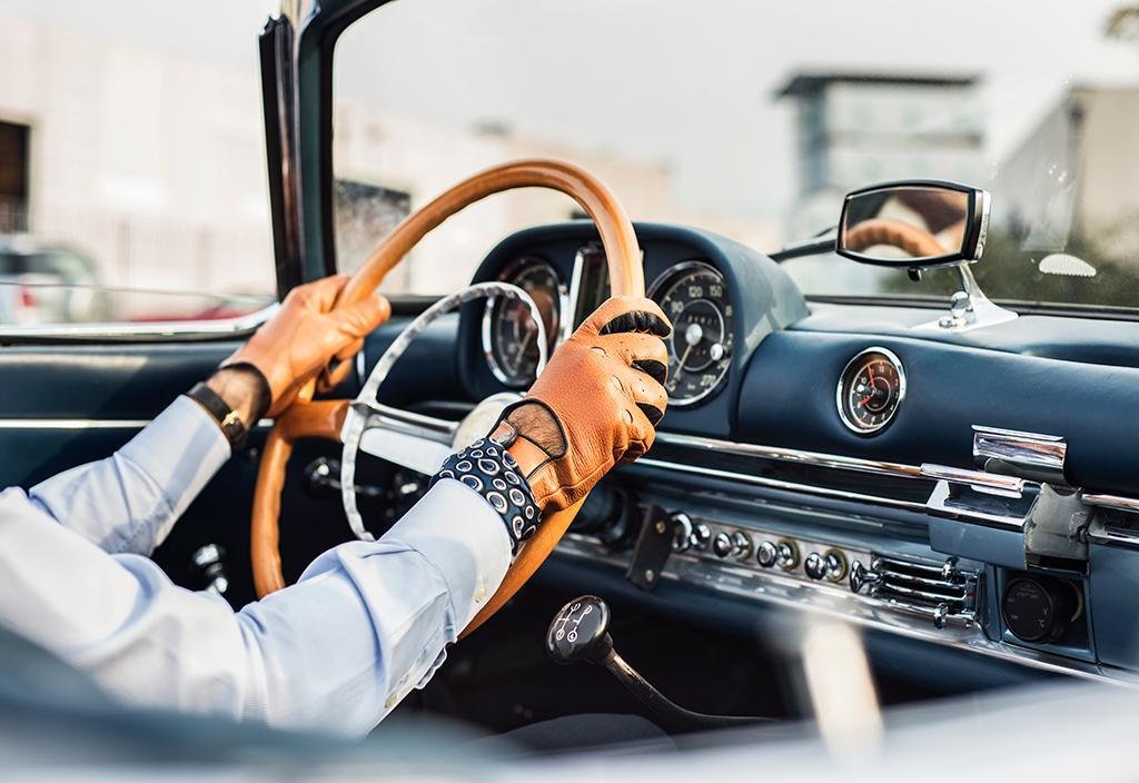 A shot of a mans hands on a luxury car steering wheel and he's wearing light brown leather gloves, the interior of the car is black and he's wearing a light blue shirt