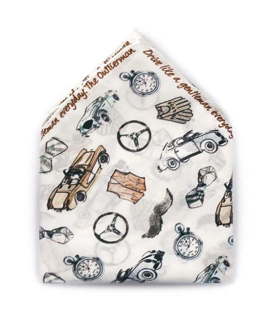 A product image of a folded white pocket square with a vintage pattern consisting of steering wheels, cars, pocket watches and cars in a vintage sketched style. The top corners have text going across them in brown handwritten font, saying "The Outlierman, Drive like a gentleman everyday"