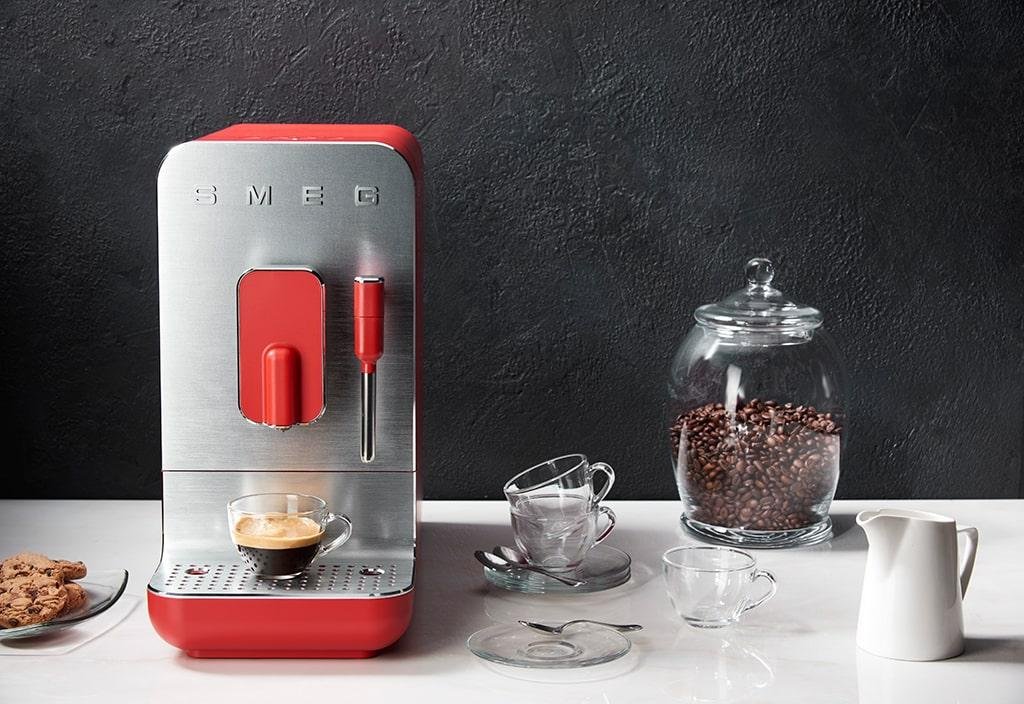 The red version with an espresso made, on a white countertop with empty cups, a jar of coffee beans and a clear plate of cookies and a jug of milk