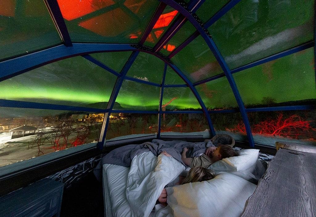 Two people laying in a bed facing the northern lights, looking out of the glass roof and admiring the view