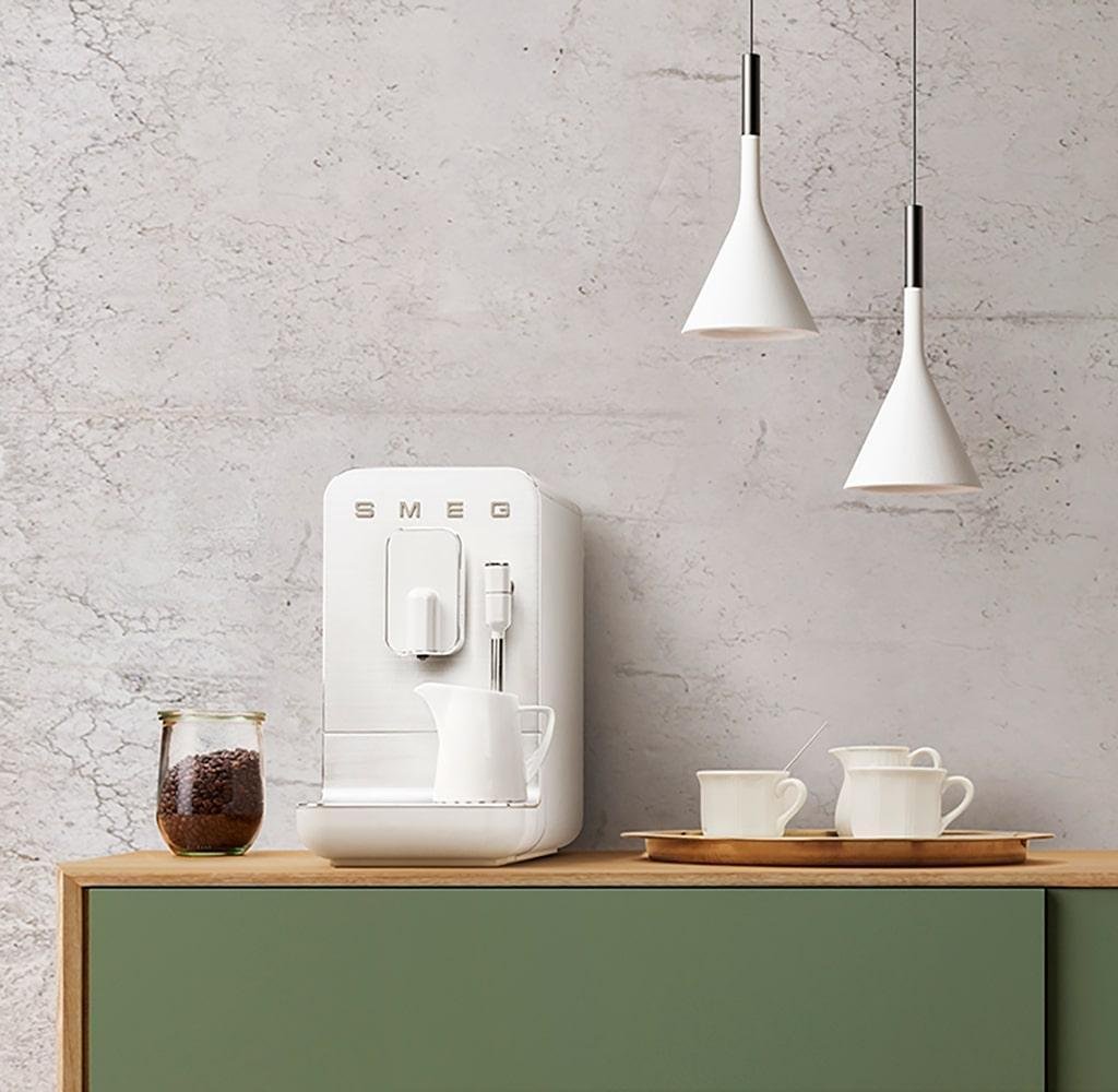 The white version with a jar of milk on it, on a wooden countertop with empty cups, a jar of coffee beans and a bowl of sugar cubes