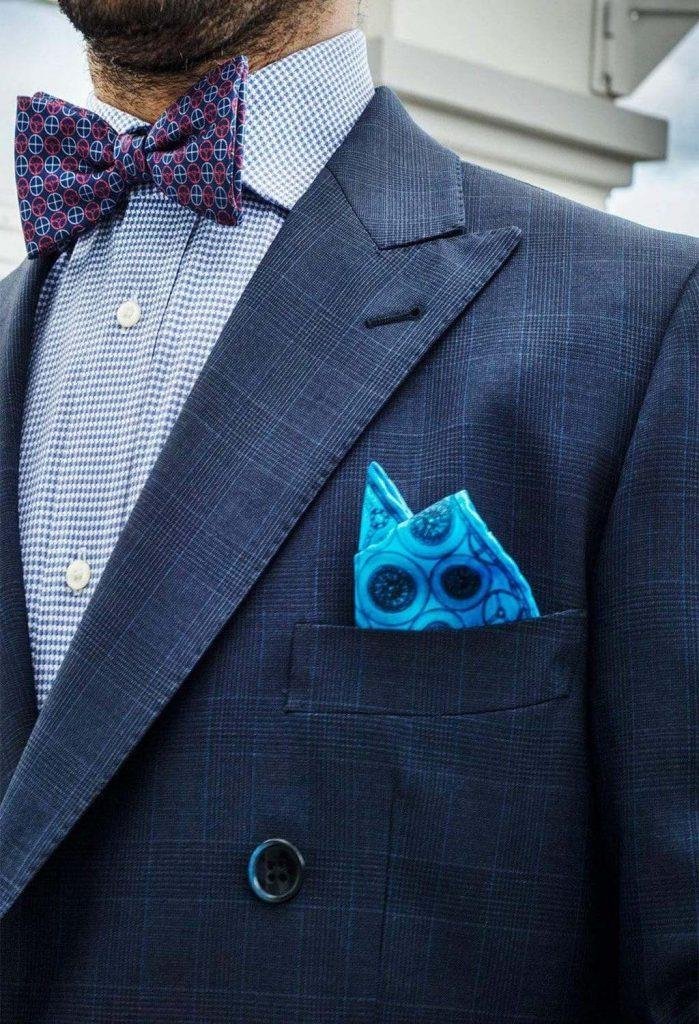 A close up of a mans navy suit with a light blue button up underneath and wearing a purple and blue patterned bow, the image focuses on a blue pocket square with a circular dark blue pattern