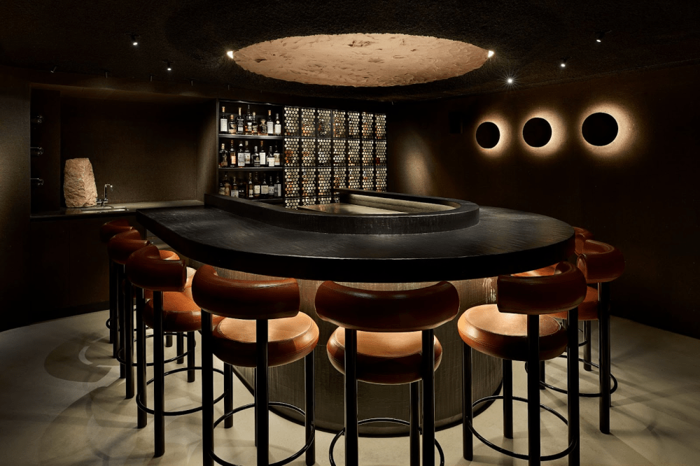 Dimly lit room, with fine mahogany bar chairs, surrounding an arch shaped smooth black table top. The back walls of the room cradling exotic whisky.